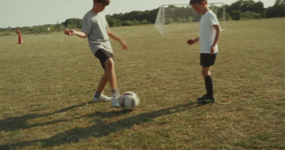 Two young boys dressed in soccer attire practice dribbling a ball on a sunny dry grassy field one boy demonstrates control over the ball with his feet while the other watches with a soccer goal visible in the background under the clear sky