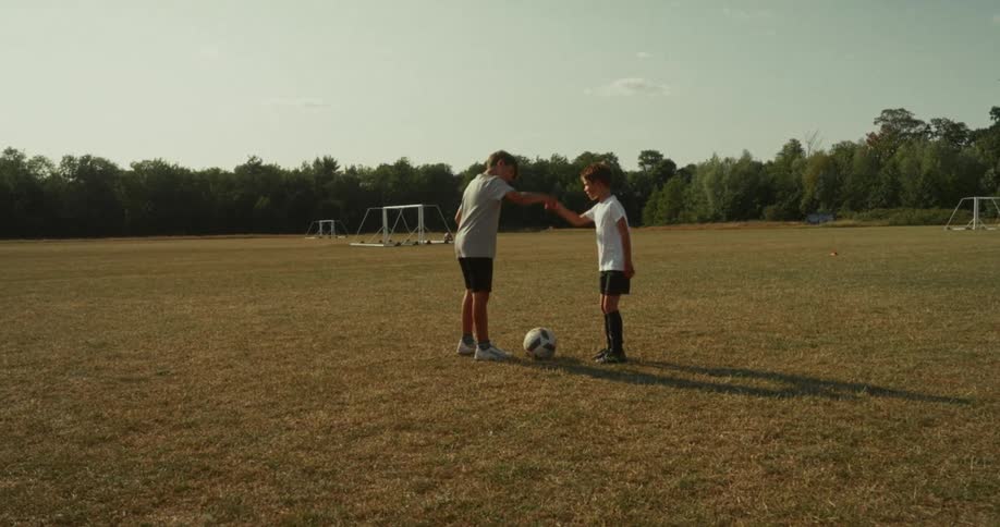Two young boys in soccer gear stand on a dry grassy field with a soccer ball positioned between them they briefly shake hands displaying good sportsmanship against a backdrop of trees and a distant soccer goal under the bright sky