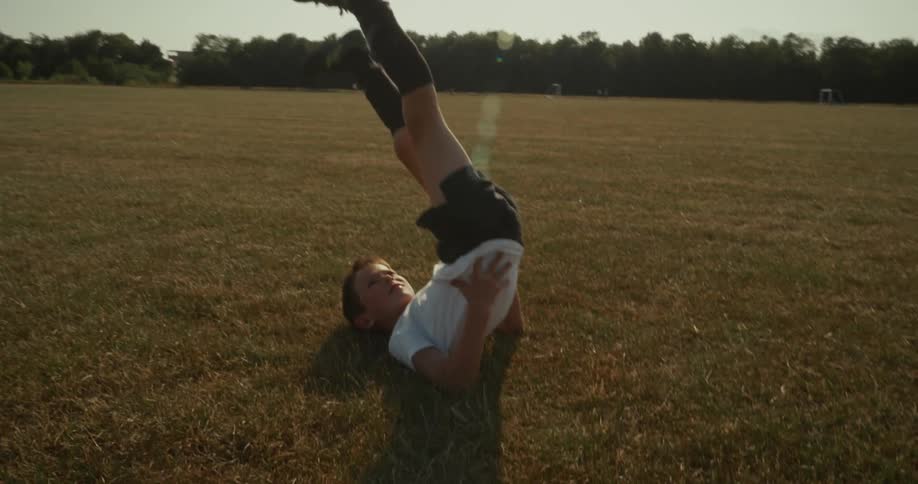 A young boy in athletic wear performs a leg raise exercise on a grassy field with his upper body on the ground and his legs extended vertically he lifts and lowers his legs in a controlled manner showcasing his core strength during