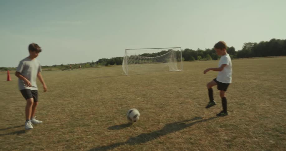 Two children kick a soccer ball back and forth on a grassy field taking turns while a goalpost stands in the background