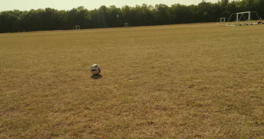 A soccer ball bounces alone across a dry grass field with a distant goalpost visible in the background