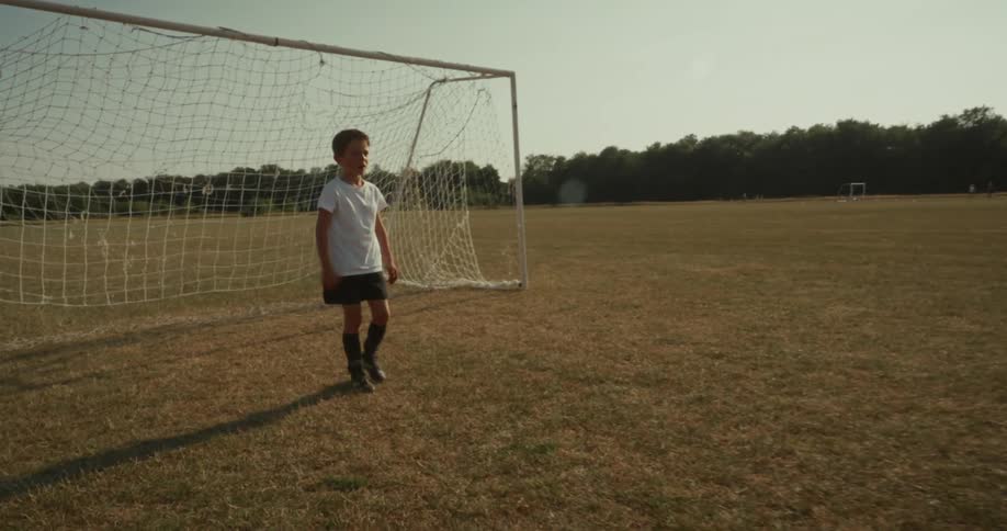A boy kicks a soccer ball on a grassy field toward a goalpost in the distance