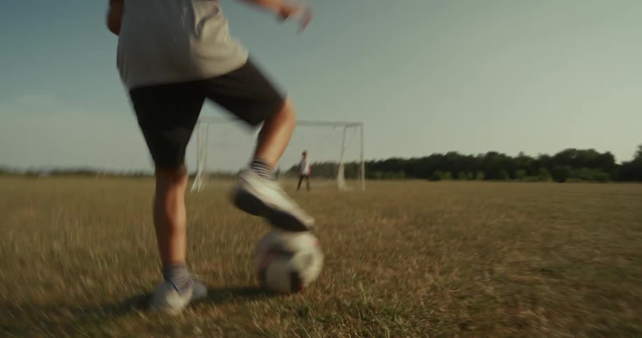 A lowangle shot follows a soccer players legs as they skillfully dribble a ball across a dry grassy field towards a goal in the background another player stands near the goal presumably the goalkeeper under the bright sunlight
