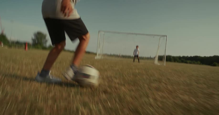 A lowangle shot of a persons legs wearing shorts and sneakers running across a grassy field towards a soccer goal the person then kicks a soccer ball and another person possibly a goalkeeper is seen in the distance near the goal