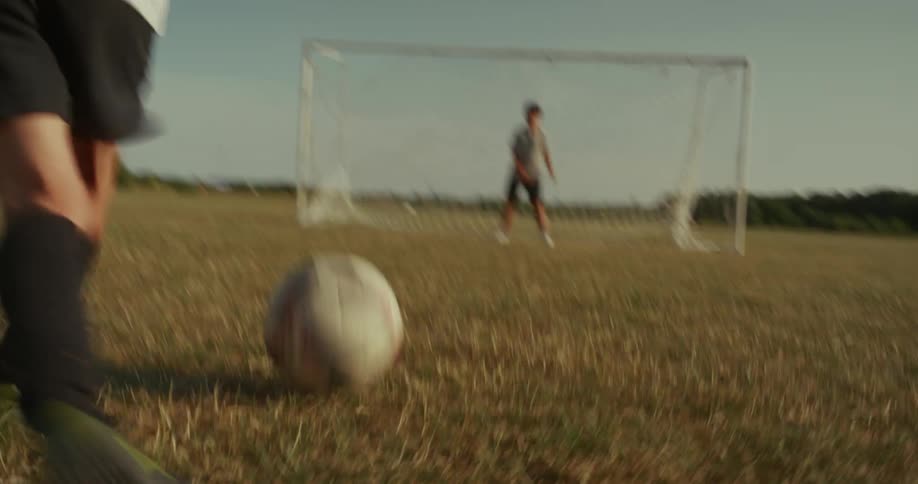 A boy kicks a soccer ball on a grassy field toward the goal where another child stands ready as the goalie