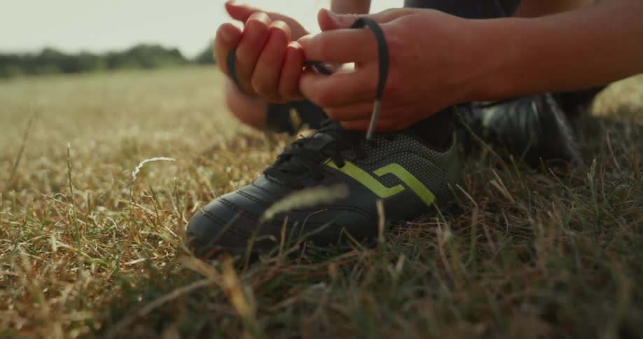 A child is sitting on a grassy field tying the laces of black soccer cleats