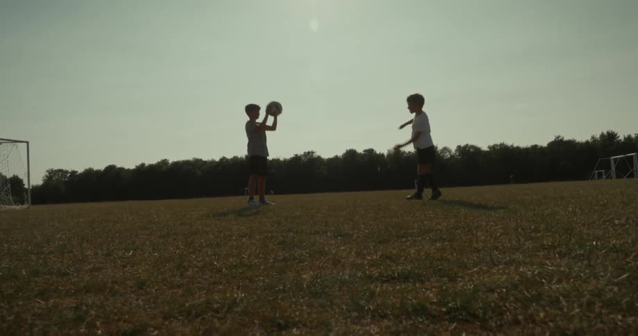 Two kids are tossing a football to each other on a soccer field at sunset