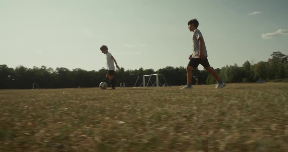 Two children play soccer on a dry grassy field with multiple goalposts in the background