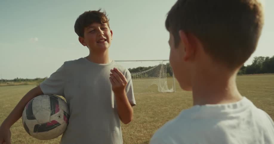 Two boys stand on a soccer field with one holding a ball and talking to the other who is facing away from the camera