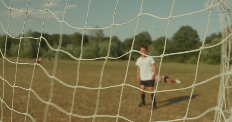A boy kicks a soccer ball across a dry grass field into the net seen from behind the goal