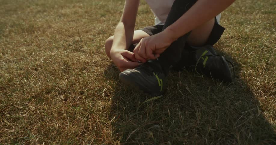 A boy sits on a dry grass field diligently tying the laces of his soccer cleats
