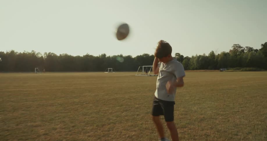 A boy on a sunny grassy field presumably a sports field with goalposts visible in the background he is looking up and appears to be in the process of catching or throwing a rugby ball which is suspended in the air above him