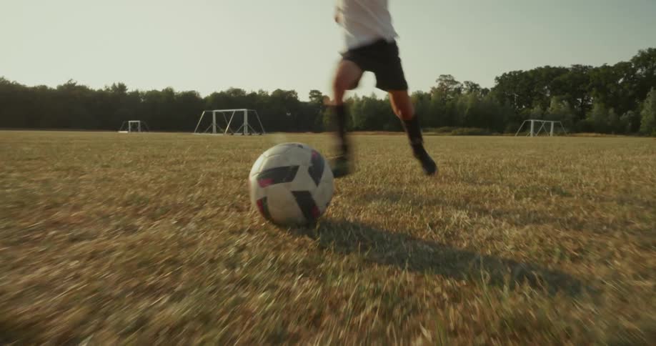 A child runs toward a soccer ball on a dry field and kicks it into a goalpost