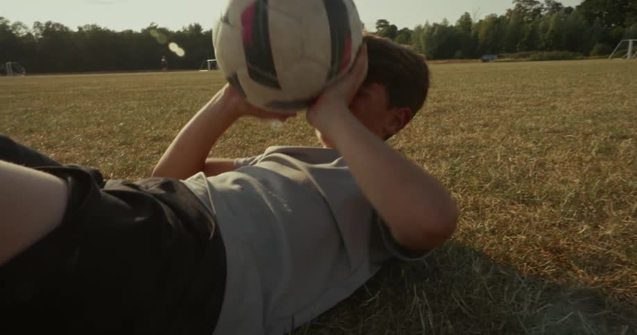 A young boy performing situps on a dry grassy field with distant goalposts suggesting a sports ground setting he is using a soccer ball as part of his exercise raising it above his head during the movement