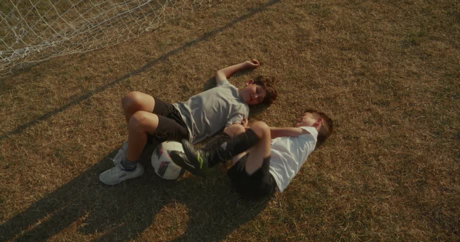 Two young boys likely soccer players lying on a dry grassy field next to a soccer ball they are wearing athletic shorts and tshirts and the background features a blurred soccer goal