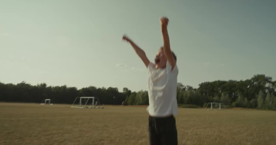 A child in a white shirt and black shorts raises both arms in celebration on a grassy soccer field with goalposts and trees in the background