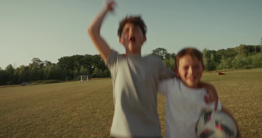 Two young boys on a dry grassy field with one boy holding a soccer ball they are both excitedly jumping and cheering with the other boy raising his arms in triumph suggesting they just scored a goal or celebrated a victory