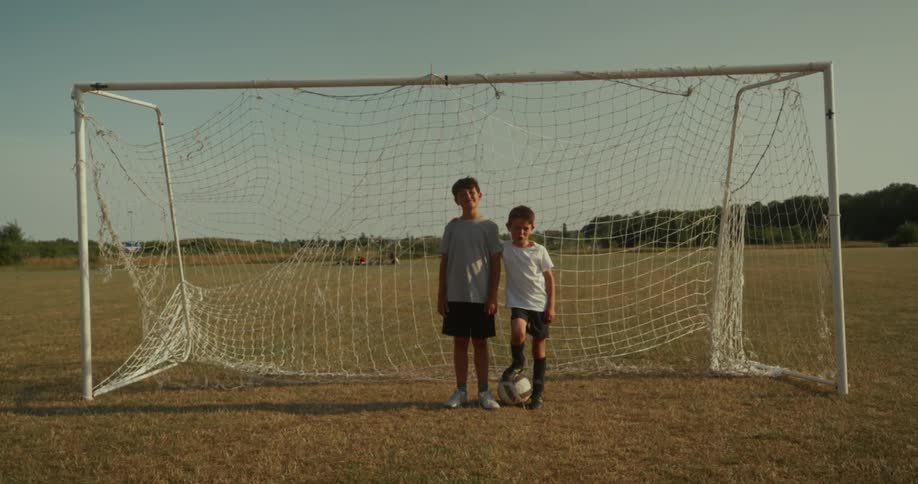 Two young boys in soccer attire standing in front of a soccer goal on a dry grassy field they are looking directly at the camera with one boy resting his foot on a soccer ball