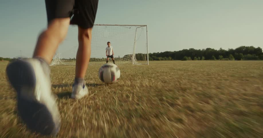 A child kicks a soccer ball toward the goal while another child stands ready as goalkeeper on a sunlit dry field