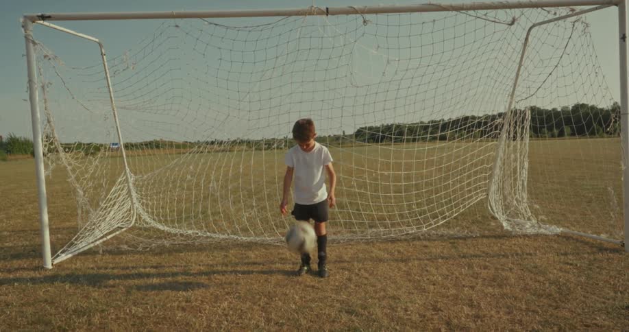 A young child stands in front of a soccer goal on a grassy field bouncing a soccer ball