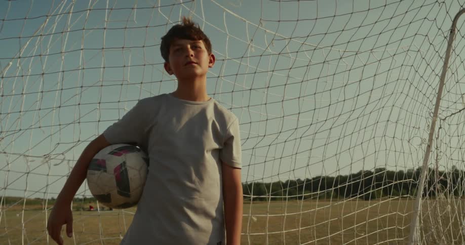 A young boy likely a soccer player standing in front of a soccer goal on a dry grassy field holding a soccer ball he looks directly at the camera with a determined or contemplative expression