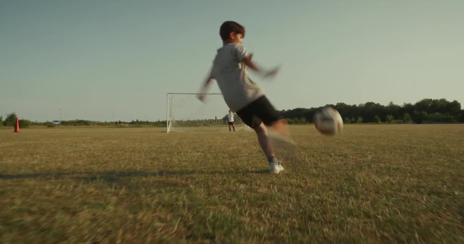 A young boy playing soccer on a vast dry grassy field under a clear sky he dribbles the ball forward and then takes a shot towards a goal where another boy appears to be acting as the goalkeeper in the distance