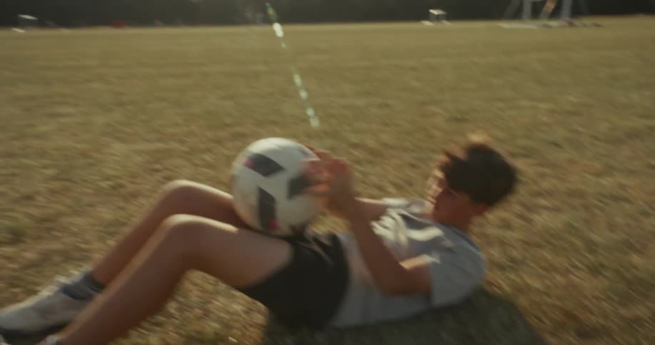 A boy engaging in a workout on a grassy field with goalposts visible in the background suggesting a sports setting he is performing exercises that involve holding and manipulating a soccer ball alternating between an upright position and lying