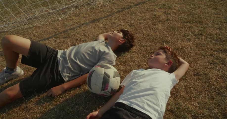 Two children lay in the grass next to a soccer ball and net