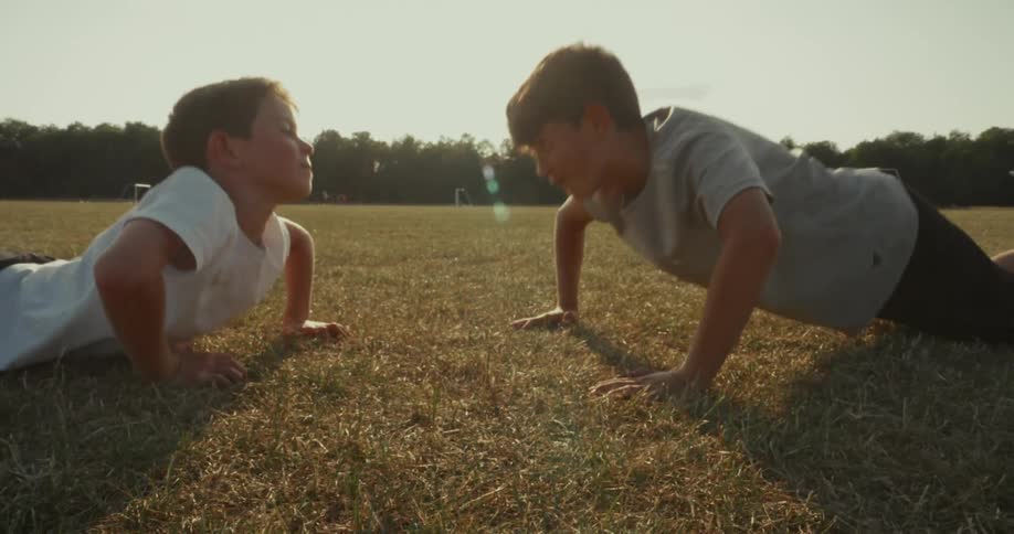 Two boys do pushups and touch hands in the dry field under the evening light