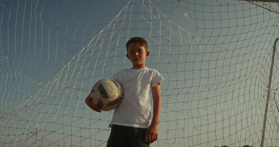 A young boy likely a soccer player standing in front of a soccer goal holding a soccer ball he looks directly at the camera with a confident expression before looking up and then back at the camera