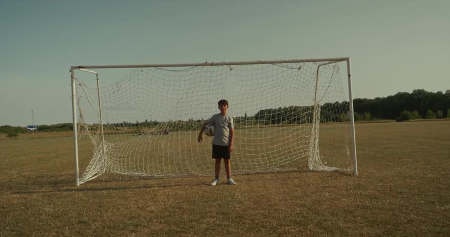 A young child stands in front of a soccer goal on a grassy field holding a soccer ball