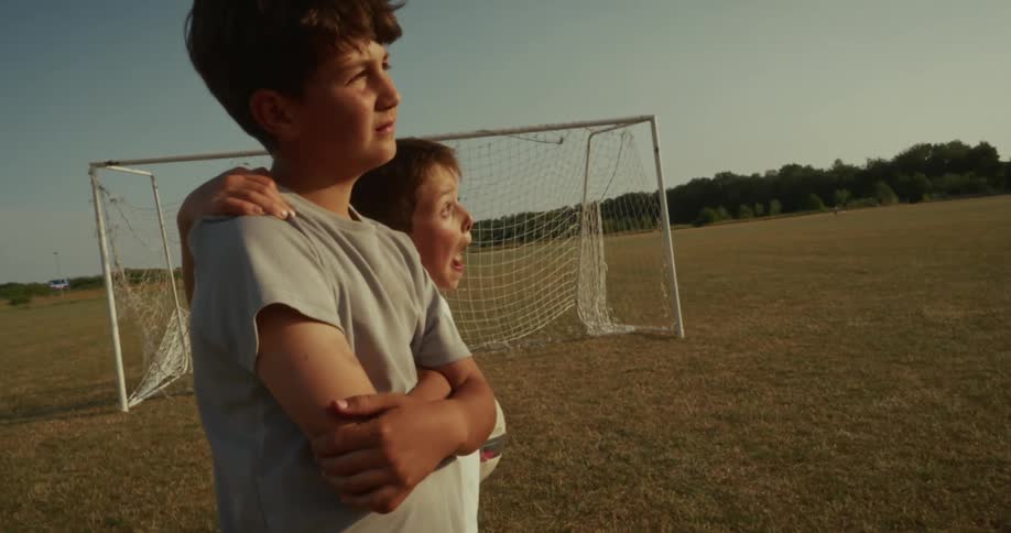 Two children stand on a sunny grassy field one holding a soccer ball with an arm over the others shoulder near a goalpost