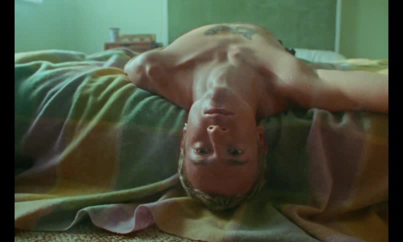 A shirtless man lies on a bed under a blanket smoking and staring upward in a dimly lit room