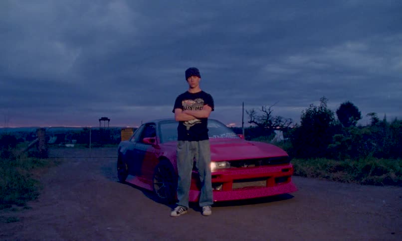 A young man with his arms crossed stands proudly in front of his custom red tuner car on a rural dirt road a single headlight cutting through the settling dusk