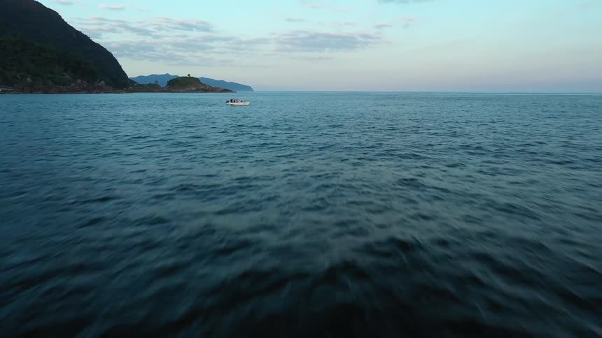 A serene aerial view of a vast expanse of dark blue ocean with small waves rippling across the surface in the distance a small white boat floats peacefully with lush green islands and mountains forming a picturesque horizon under a clear sky