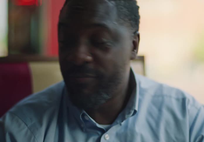 A darkskinned man with a beard wearing a light blue collared shirt is shown in a closeup he looks downwards and then looks up and chews on something