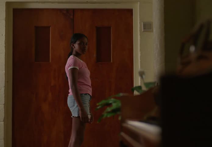 A young girl with dark hair wearing a pink tshirt and denim shorts walking through an indoor space towards a set of closed wooden double doors she glances towards the right of the frame as she passes by where a blurred green plant is visible
