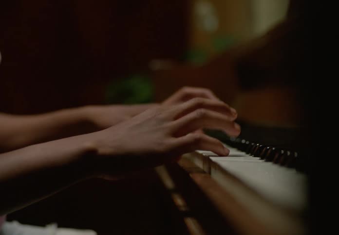 A person with dark skin and a ponytail plays the piano their hands moving across the keys in a closeup shot the video then shows a profile view of the person focused and engrossed in their playing