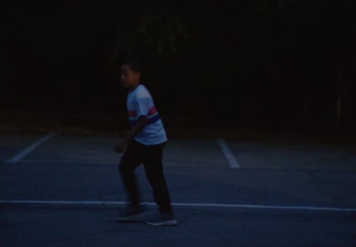 A boy in a white and blue shirt runs across a dark empty street at night he runs back and forth appearing to be playing a game or exercising with a vehicle passing in the background