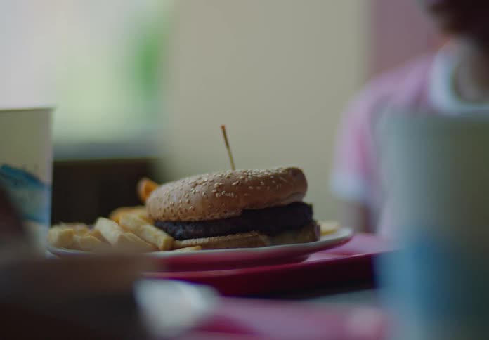 A mouthwatering closeup of a burger and fries takes center stage on a red plate the camera then pans to reveal a young girl with dark hair looking thoughtful as she sits at the table