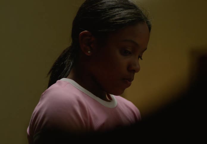 A young girl with dark hair tied in a ponytail is shown in a closeup looking downwards with a thoughtful expression she is wearing a light pink tshirt with white trim and the lighting is dim suggesting an indoor or lowlight setting