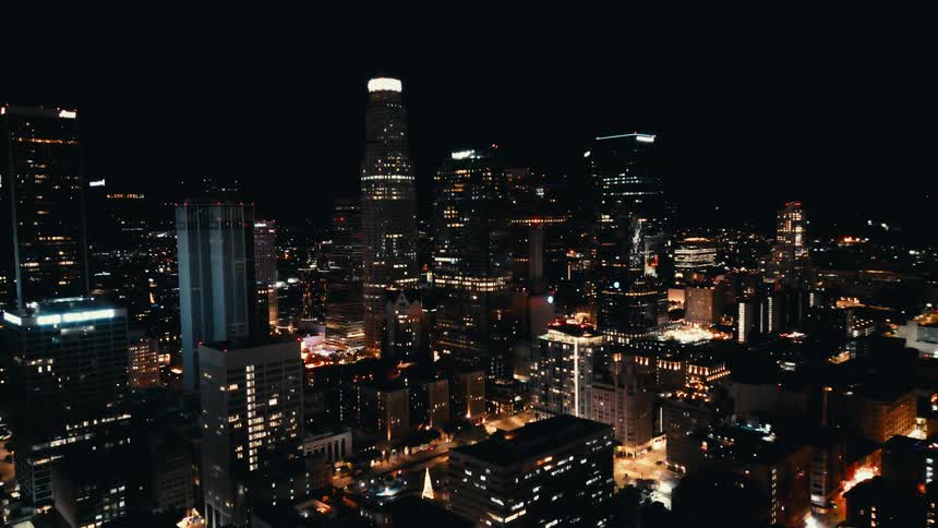 A nighttime aerial view of the city shows numerous buildings and skyscrapers lit up against the dark sky
