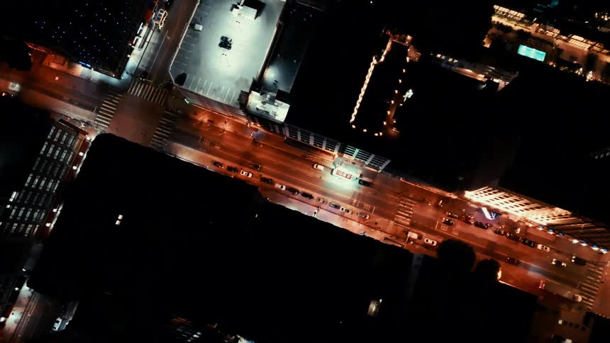 An aerial drone shot captures a city street intersection at night with car headlights and building lights creating a warm glow against the dark urban landscape