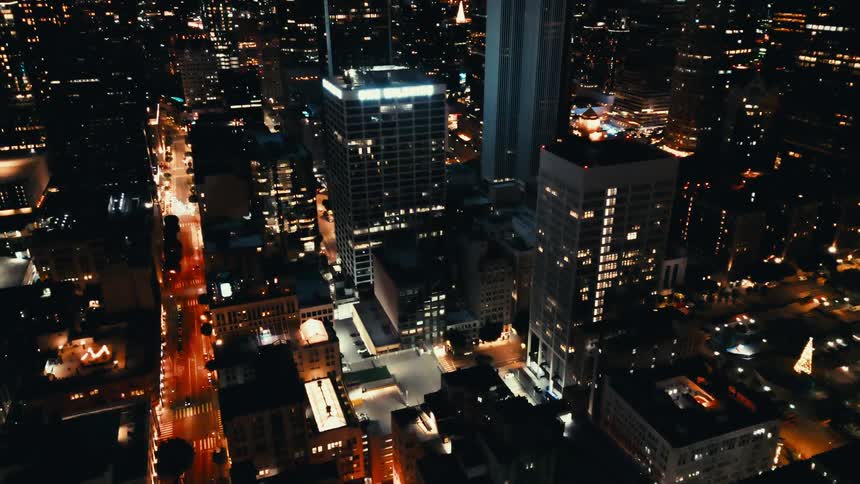 City streets glow under bright lights flanked by towering highrise buildings against a night sky
