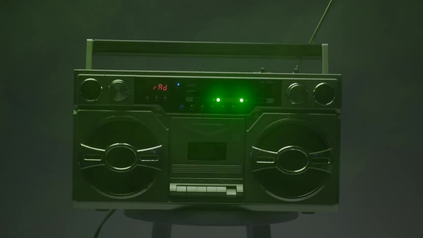 A vintage boombox with illuminated buttons and dials is positioned against a dark background under flashing colorful lights