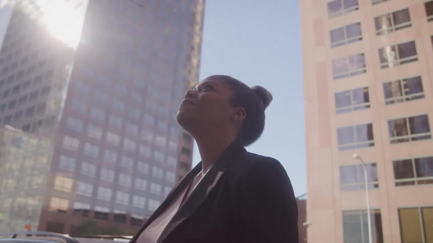 A woman in business attire stands in front of tall buildings as sunlight reflects off the glass faade