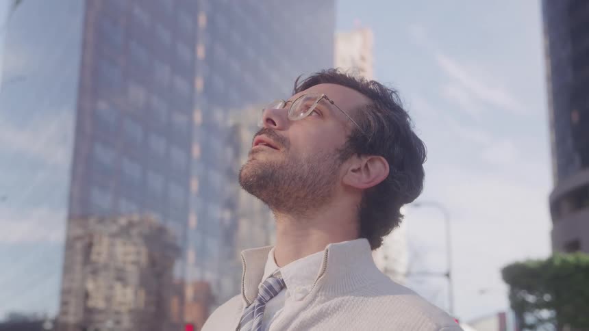 A man wearing glasses and a striped tie looks up at tall city buildings under clear sunny skies