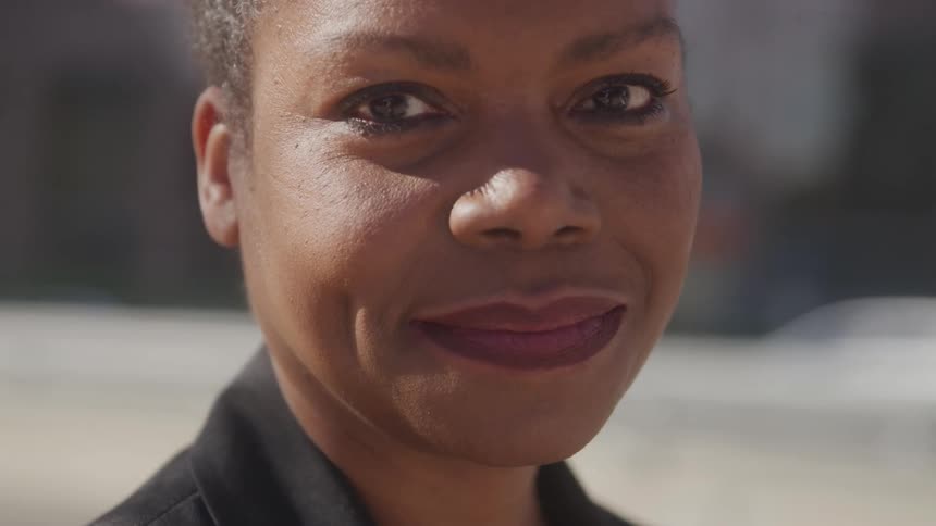 A woman in a black blazer and white shirt stands smiling directly at the camera