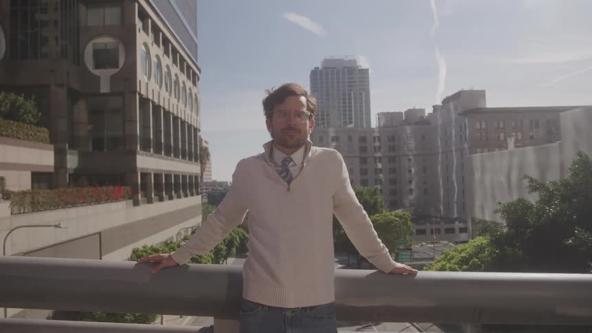 A person stands on a bridge railing above a city street surrounded by tall buildings and leafy trees