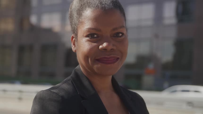 A woman in a black blazer and white shirt stands in front of a multistory building smiling directly at the camera